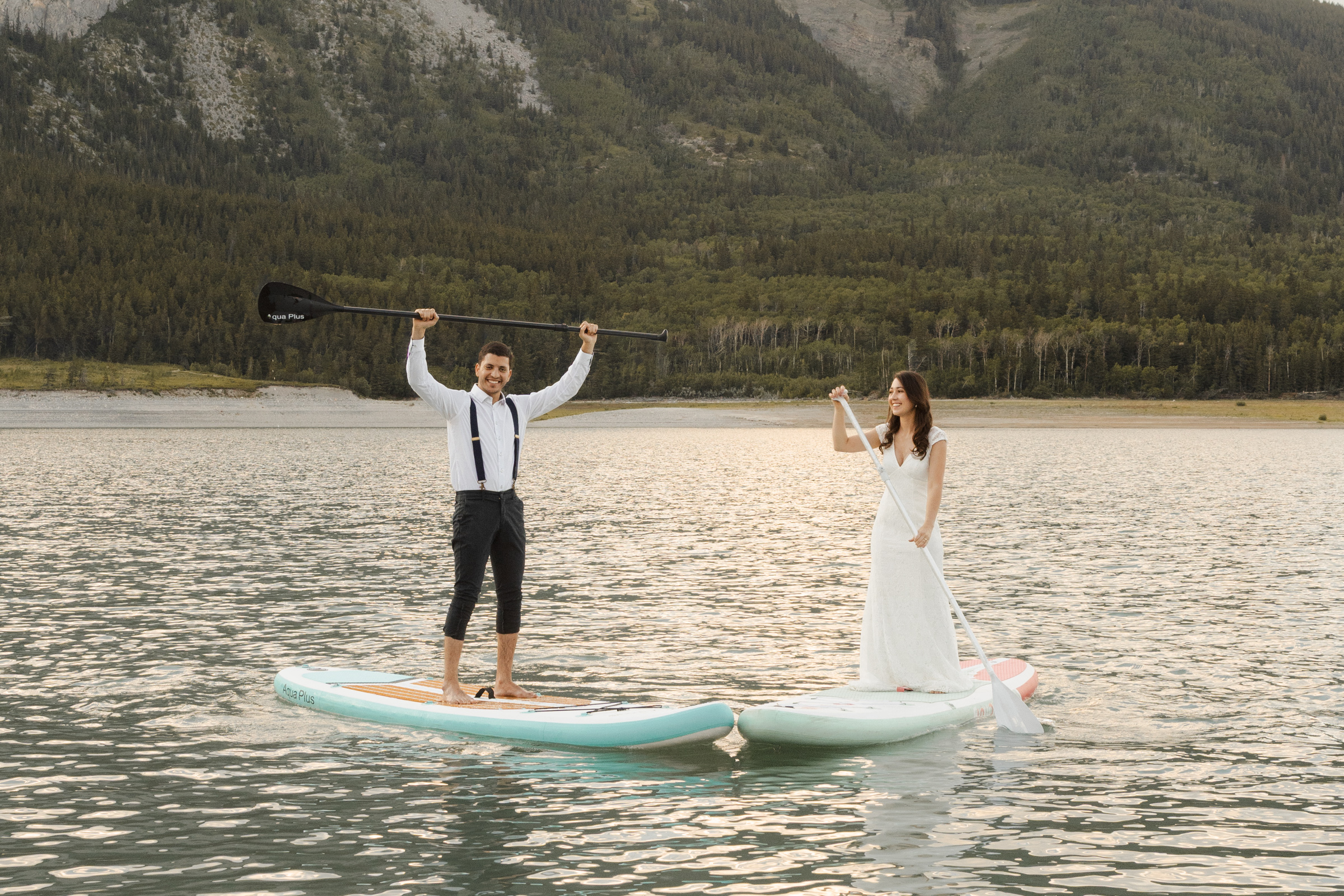 A bride and groom in wedding attire stand on paddleboards on the calm water of Spray Lakes in the Canadian Rockies, with mountains in the background.